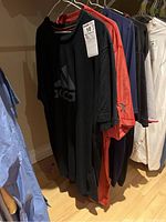 Photo showing five men's sports shirts hanging on a clothing rack, includes a black Adidas shirt with visible tag, and other colored shirts including red and navy.