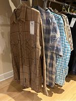 Six men's button-up shirts hanging on hangers, showing various plaid and check patterns in earth and blue tones.