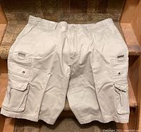 Beige RRJ cargo shorts laid flat on a brown carpeted stair, multiple side pockets visible, belt loops, and zipper fly.