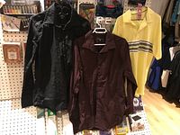 Three men's tops hanging on black and white plastic hangers against a pegboard wall. Tops include a black long sleeve button-up shirt, a maroon long sleeve button-up shirt, and a yellow polo shirt with multiple horizontal stripes.