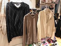 Three men's sweaters hanging on white hangers against pegboard wall: Tommy Hilfiger black argyle, Mexx beige V-neck, Guess cream zippered sweater.