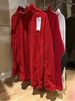 Three men's long-sleeved tops hanging on hangers, including two reds fleece tops and one beige turtleneck shirt.