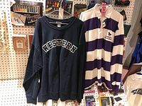Two men's shirts hanging on display: a navy blue 'WESTERN' sweatshirt and a purple-white striped rugby shirt with collar and logo.