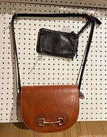 Tan Argentinian leather purse hanging on pegboard with gold horse bit detail and long black adjustable strap, shown with black leather Roots wristlet attached.