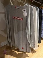 Row of five men's long-sleeved shirts on hangers including Roots grey 'CANADA' shirt, white, light blue, striped blue and dark grey shirts