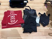 Top view of red Nike Pro leggings, black Nike long underwear leggings, grey and black New Balance leggings, black Nabaiji sports bra, folded clothes and scarves on wooden floor.