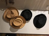 Four hats on pegboard including two natural straw colored hats and two black colored hats, displayed hanging.