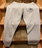 Full view of grey sweatpants with Sneaktip logo on left thigh, showing elastic waistband and ankle cuffs.