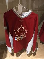 Full view of red Roots Sydney 2000 Olympic jersey showing front design, maple leaf with torch and Olympic rings, hanging on white hanger.