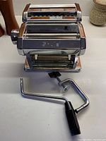 Full view of chrome Shule pasta maker showing the body, rollers, and detached handle and clamp attachments.