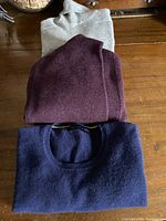 Photo showing folded light grey turtleneck, dark burgundy sweater, and dark blue sweater stacked on wooden surface, front view.