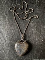 Photo of the heart-shaped silver locket pendant with detailed floral etching on a dark background showing front side.
