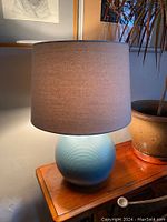 The full lamp with the blue ceramic base and the beige linen shade on a wooden table next to a potted plant.