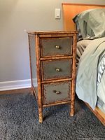 Full view of one bamboo nightstand with three drawers, showing overall shape and size relative to bed and carpet.