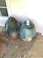 Two large ceramic or terra cotta pots with molded lids set outside on stone floor, showing exterior surface and shape.