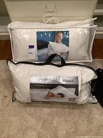 Two pillows in original packaging: one Cuddledown down pillow in striped fabric zip bag, one Seasons Pinnacle King allergy-free down pillow in clear bag with black trim