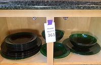 View of stacked large and smaller dark emerald green glass plates and bowls arranged on a wooden shelf.