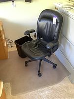 Full view of the black leather office chair placed on clear floor protector mat with black wastebasket beside it.
