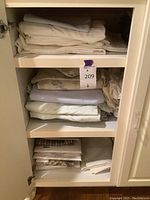 Full view of linen stacks on three shelves inside a white cabinet, showing folded sheets and pillowcases in neutral tones and some in original packaging.