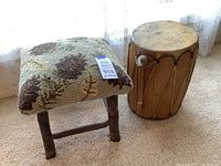 Rustic wooden stool with green and brown autumnal hooked seat and a wooden leather drum side by side on carpeted floor.
