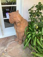 Right side view of terra cotta tiger statue placed against a white door and stone floor with plants nearby.