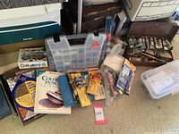 Overview of assorted art supplies including books, plastic organizer box, packs of art tools and paint tubes on floor