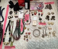 Wide shot of assorted jewelry pieces laid out on a white quilted fabric showing necklaces, earrings, brooches, and pins with pink labels identifying Murano glass, Jade, and IOU semi-precious stones.