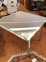 White folding table with a rectangular plastic top and gray metal folding legs, placed on a patterned rug indoors.