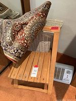 Wooden folding chair with slatted seat next to white wicker hamper and compact space heater on carpeted floor. Persian rug partially draped over chair.