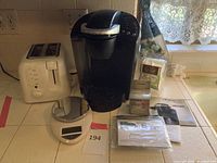 Photo showing Keurig coffee maker, white toaster, digital kitchen scale, and Keurig coffee supplies on kitchen counter