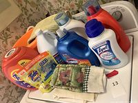 Photo showing various laundry detergent bottles (red, blue, white), Tide PODS box, Lysol Laundry Sanitizer bottle, and decorative towel with farm print laid on top in front of washing machine.