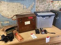 Old microscope in wooden case along with binoculars, golf scope, briefcase and file box arranged on a table.