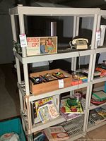 Plastic shelving unit with four shelves holding various items for size reference, includes game box, telephone, and books.