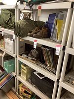 Wide angle photo showing white plastic shelving unit with various items stored on it such as bags, plastic boxes, books, and miscellaneous items. Shelving unit has horizontal slatted shelves and tubular frame.