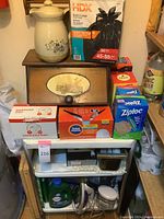 Wooden shelf with drawer containing kitchen and cleaning items including ceramic breadbasket, trash bags, Ziploc bags, iron, and detergent.