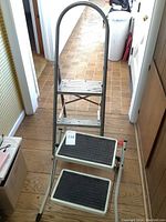 Metal step ladder with two steps and a handle, shown standing on wooden floor in hallway.