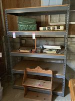 Full frontal image of the metal shelving unit showing four shelves, various items on shelves including a green decorative box, books, and papers. Shelving stands on concrete floor against wood paneled wall.