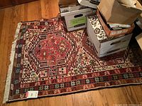 Full view of rectangular wool rug laid on wooden floor surrounded by boxes, showing rich geometric patterns and fringed edges.