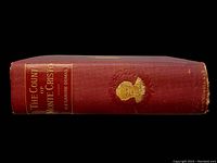 Side view of the book's spine showing the title, author's name, and a gold embossed portrait on textured red cloth cover.