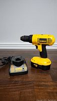 Photo showing side view of yellow and black DeWalt cordless drill with battery attached and charger next to it on a wooden surface.