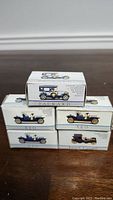 Five small vintage car model boxes stacked in two layers on a wooden surface showing the illustrations and branding of the cars.