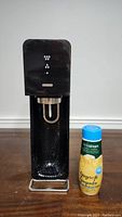 Full view of black Sodastream soda maker showing carbonation head and front markings, with Ginger Ale flavor syrup bottle to the right side