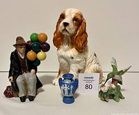Photo showing four figurines: balloon seller man holding multi-colored balloons, sitting Spaniel dog, small blue and white vase, and hummingbird on flowers figurine.