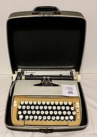 Front view of white and beige vintage typewriter with round keys inside open black carrying case with handle.