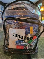 Front view of the clear backpack showing the Amazon logo, staples stapler, scissors, white glue, and assorted folders and notebooks inside.