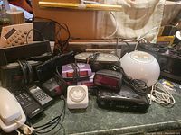 Wide shot showing an assortment of black and white used electronic devices including remote controls, analog and digital clocks, telephones, and timers scattered on a dark countertop.