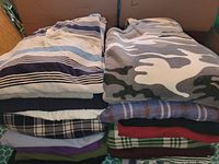 View of stacked folded men's tops showing fabrics and patterns including stripes, camouflage, plaid and solids