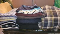 Stack of folded men's clothing items including checkered, plaid shirts and solid tops, showing variety of patterns and fabric textures