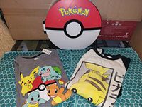 Photo showing two Pokemon-themed t-shirts and a round Pokemon-themed storage case from the front