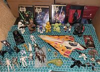 Wide shot of Star Wars DVDs standing behind a collection of approximately 30 Star Wars character figurines and a toy fighter ship in front. Figurines include main characters Darth Vader, Yoda, Grogu, R2-D2, C-3PO, stormtroopers, and others. DVDs displayed in their original cases.